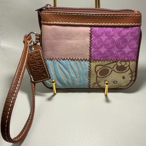 Puede incluir: Un monedero de patchwork con una correa de muñeca de cuero marrón. El bolso presenta una variedad de telas, incluyendo rosa, azul y morado, con un gráfico de Hello Kitty. El bolso está sostenido por dos soportes dorados.