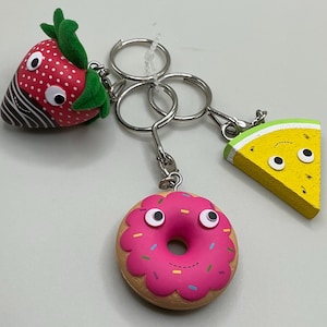 May include: A keychain with three charms: a red strawberry with green leaves, a pink donut with sprinkles, and a yellow watermelon slice. Each charm has a cartoon face. The charms are attached to a silver metal keyring.