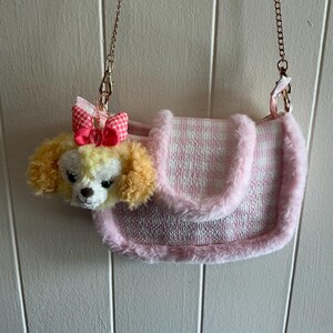 May include: A pink and white patterned handbag with a plush, fluffy trim. The bag features a dog-shaped embellishment with a pink bow and a gold chain strap. The dog has yellow and white fur.