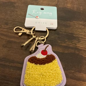 May include: A novelty keychain featuring a dessert design. The keychain has a yellow, textured base, topped with brown and white accents, and a red cherry. The tag reads "Key ring" and "OMISE". The keychain has a gold-colored clasp.