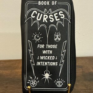 May include: Black zippered wallet with white graphic text and illustrations. The text reads "BOOK OF CURSES" and "FOR THOSE WITH WICKED INTENTIONS". The wallet has a rectangular shape and is propped up on gold-colored metal stands.