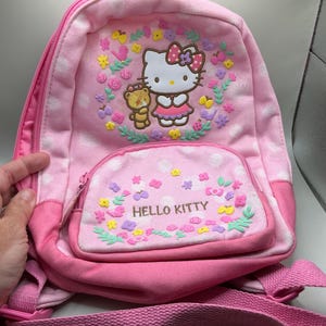 May include: Pink Hello Kitty backpack with a front pocket. The backpack features a Hello Kitty graphic with a teddy bear, surrounded by flowers. The front pocket has the words "Hello Kitty" printed on it. The backpack has a pink shoulder strap and a zipper closure.