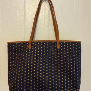 Puede incluir: Un bolso tote azul marino con un estampado de estrellas doradas. Tiene asas y ribetes de cuero marrón. El bolso rectangular está hecho de un material similar a la lona.
