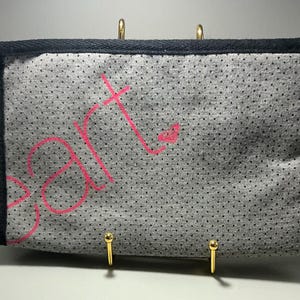 May include: Gray clutch with black trim and a black wrist strap. The clutch has a dotted pattern and the word "art" is written in pink. Gold-colored hooks are attached to the bottom.