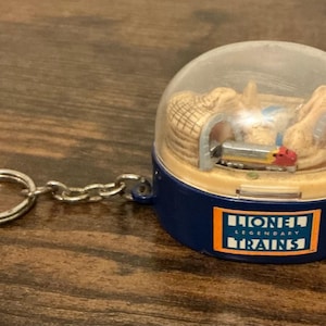 Può includere: Portachiavi blu e trasparente con una scena di treno in miniatura all'interno di una cupola. Il portachiavi ha una catena e un anello argentati. La base della cupola ha un logo Lionel Trains.