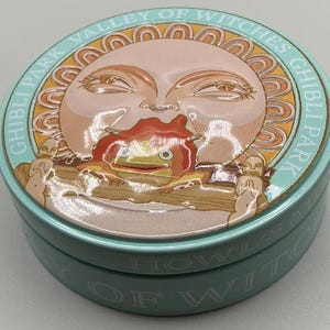 May include: A round, light blue tin with a detailed illustration. The lid features a sun-like face with a red and orange creature. The text "GHIBLI PARK VALLEY OF WITCHES" is printed around the edge. The tin is likely for storage or a collectible item.