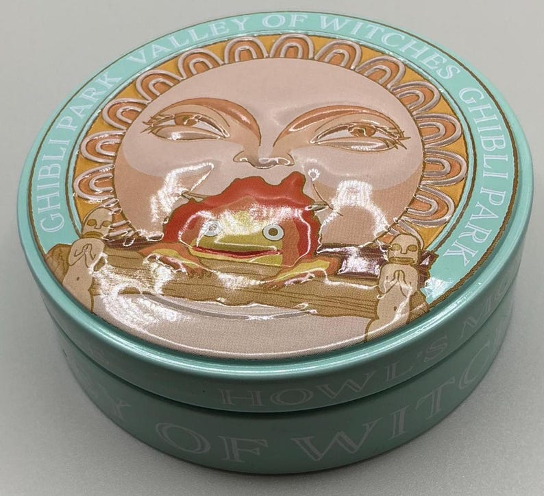May include: A round, light blue tin with a detailed illustration. The lid features a sun-like face with a red and orange creature. The text "GHIBLI PARK VALLEY OF WITCHES" is printed around the edge. The tin is likely for storage or a collectible item.