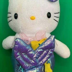 May include: White plush Hello Kitty toy dressed as a mermaid with a purple iridescent tail and a yellow star. She is wearing a pink bow.