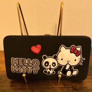 May include: Black clutch with gold-colored metal accents. The clutch features embroidered images of Hello Kitty, a panda, a red heart, and the words "HELLO KITTY" in white and gray.