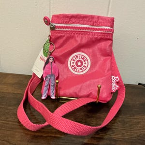 May include: A bright pink Kipling crossbody bag with a white zipper and a Barbie logo. The bag has a matching pink strap and a small Barbie doll charm attached. The bag is made of a lightweight material.