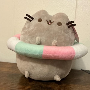 May include: A plush toy shaped like a grey cat with dark brown embroidered whiskers, eyes, and a mouth. The cat is wearing a soft, circular pool floatie around its middle, featuring alternating segments of pastel pink, mint green, and white fabric.