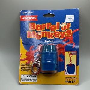 May include: A blue Barrel of Monkeys keychain on a blue and yellow card. The card reads "Barrel of Monkeys Keychain" and "Really Works!" The keychain features a blue barrel and a silver clasp. The packaging warns of choking hazards.