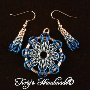 Pode incluir: Um conjunto de brincos e um colar com pingente de prata e azul. O pingente é uma peça redonda prateada com um design de malha de corrente azul e prata em forma de flor. Os brincos são de prata com um design de malha de corrente azul e prata.