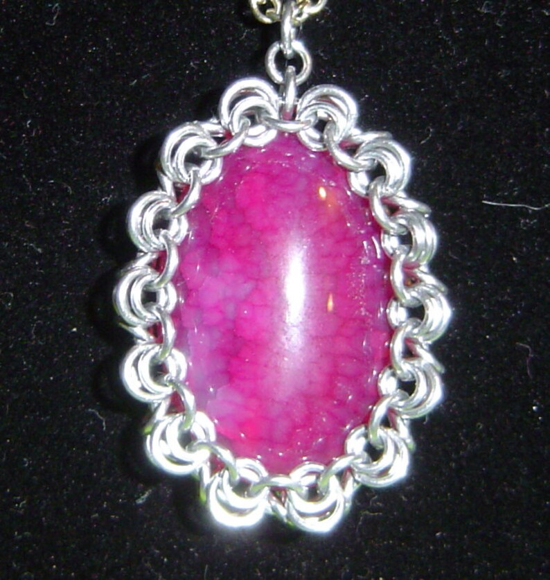 May include: A silver chainmail pendant featuring a pink agate gemstone. The pendant is oval shaped and the gemstone is set in a silver bezel.