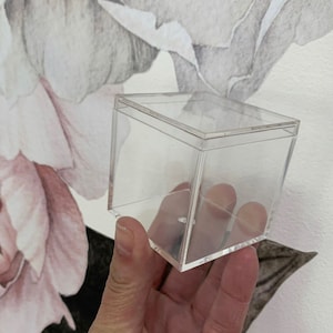 May include: A clear plastic cube-shaped box with a lid. The box is empty and appears to be made of acrylic.