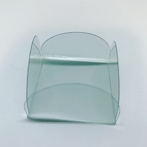 Op de afbeelding: Een transparante plastic bloempot met een vierkante basis en afgeronde randen. De pot is ontworpen om een ​​enkele bloem of een kleine plant te bevatten.