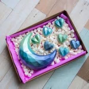 May include: A decorative gift box with a pink interior, filled with white packing peanuts. The box contains a large, faceted crescent moon and several heart-shaped objects in shades of blue and green.
