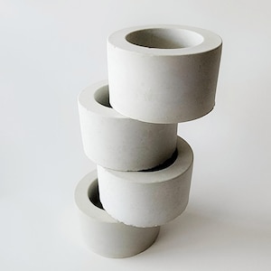 May include: Four stacked, cylindrical, light gray concrete candle holders. Each holder has a circular opening at the top and a slightly tapered base. The holders are arranged vertically against a white background.