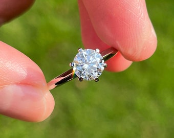 Puede incluir: Un anillo de compromiso de plata con un diamante redondo y transparente.