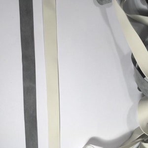 May include: Two strips of elastic, one white and one gray, are laid out on a white surface. A pile of the same white elastic is in the lower right corner of the image.
