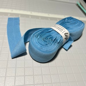 Peut inclure: Un rouleau de tissu &eacute;lastique bleu clair avec une &eacute;tiquette blanche indiquant "uip FASHION SUPPLIES" sur une surface textur&eacute;e grise.