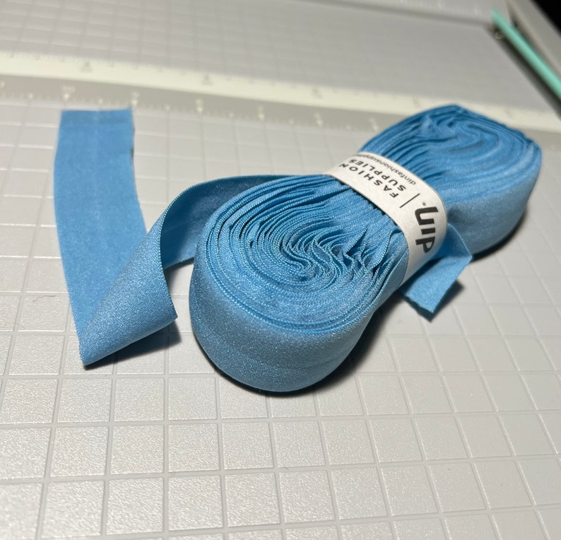 Peut inclure: Un rouleau de tissu &eacute;lastique bleu clair avec une &eacute;tiquette blanche indiquant "uip FASHION SUPPLIES" sur une surface textur&eacute;e grise.
