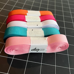 May include: Five spools of ribbon in various colors, including orange, pink, white, teal, and light pink. The spools are stacked on top of each other and have a white label with the text "dip FASHION SUPPLIES".