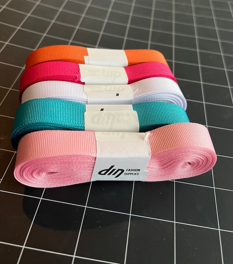 May include: Five spools of ribbon in various colors, including orange, pink, white, teal, and light pink. The spools are stacked on top of each other and have a white label with the text "dip FASHION SUPPLIES".