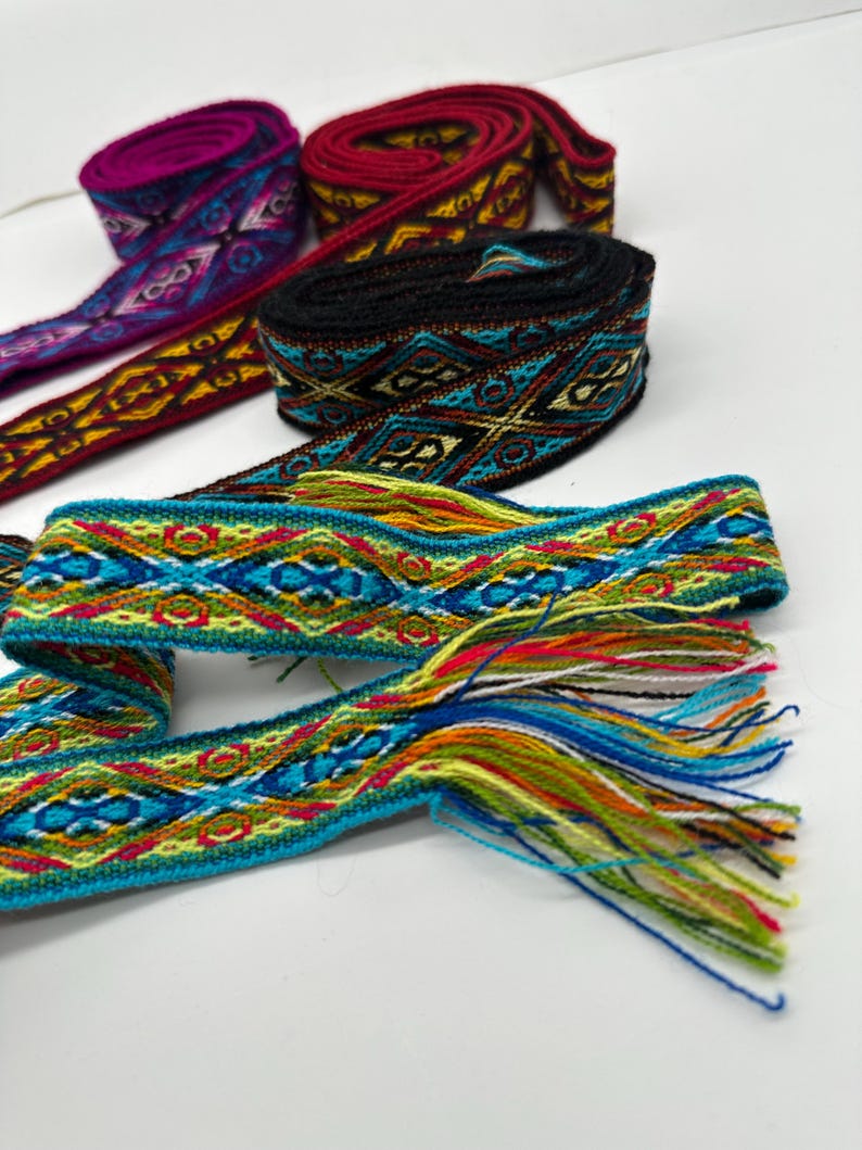 May include: Five colorful woven fabric ribbon trims with geometric patterns. The trims are in shades of red, black, blue, green, yellow, and purple. The trim on the bottom has a fringe.