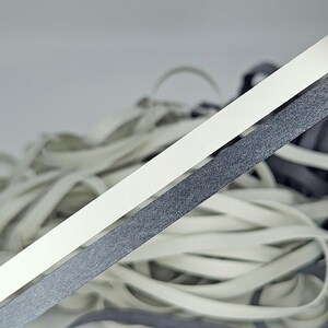 May include: Two strands of elastic cord, one white and one black, are shown against a white background. The cords are lying on top of a pile of other white elastic cords.
