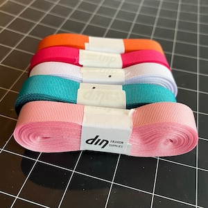 May include: A stack of five different colored satin ribbon rolls. The rolls are pink, teal, white, red, and orange. The ribbon is wrapped around a white cardboard core with a white label that says "dip FASHION SUPPLIES".