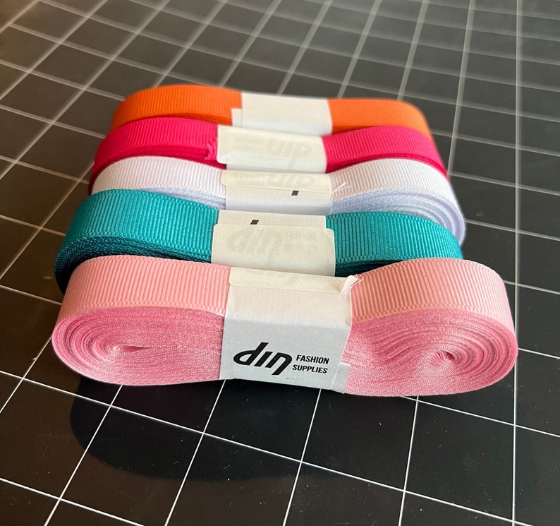 May include: A stack of five different colored satin ribbon rolls. The rolls are pink, teal, white, red, and orange. The ribbon is wrapped around a white cardboard core with a white label that says "dip FASHION SUPPLIES".