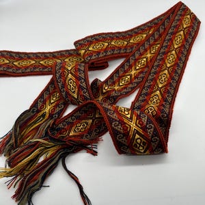 May include: A woven red, gold and blue ribbon with a geometric pattern. The ribbon has a fringe on one end.