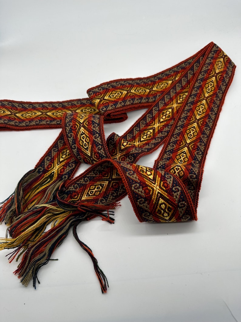 May include: A woven red, gold and blue ribbon with a geometric pattern. The ribbon has a fringe on one end.