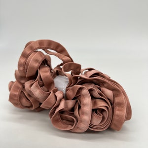 May include: A roll of dusty rose colored satin ribbon with a slight sheen. The ribbon is folded and twisted into a loose bundle.
