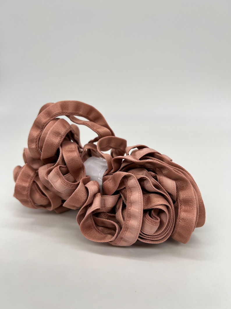 May include: A roll of dusty rose colored satin ribbon with a slight sheen. The ribbon is folded and twisted into a loose bundle.