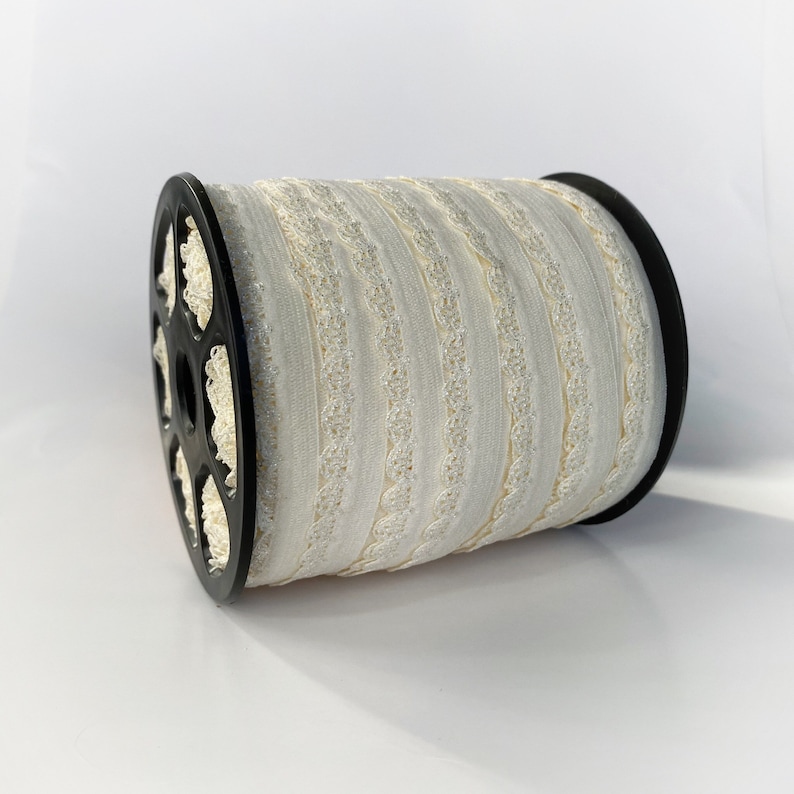 May include: A spool of white elastic trim with a silver glitter design. The trim is wound around a black plastic spool.