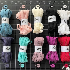 May include: A collection of colorful decorative ribbons in various shades, including teal, pink, cream, black, white, purple, red, and teal. Each ribbon is bundled and labeled with a white band featuring the text "din".