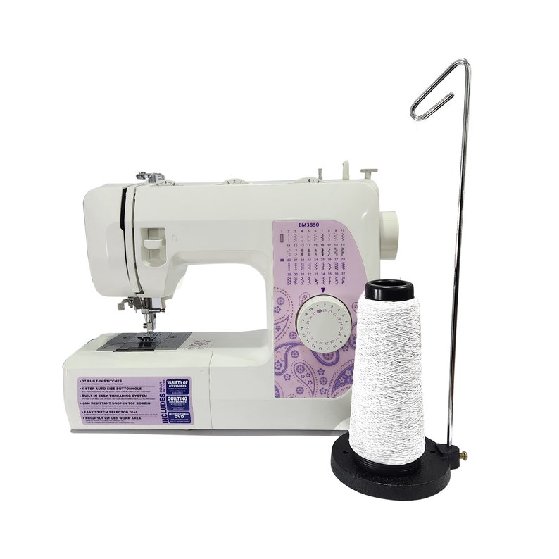 May include: White sewing machine with a purple and paisley design on the side. The machine has a built-in needle threader and a variety of stitch options. A white spool of thread is on a stand to the right of the machine.