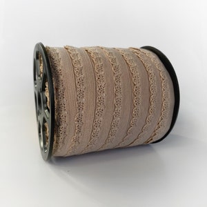 May include: A spool of beige elastic trim with a scalloped edge. The trim is wound around a black plastic spool.