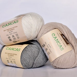 May include: Three skeins of yarn in white, beige, and gray. Each skein has a label that reads "CANCAS" and includes the yarn composition: 85% Tangus Cotton and 15% Alpaca. The label also states "Baby" and the weight, length, and yardage of the yarn.