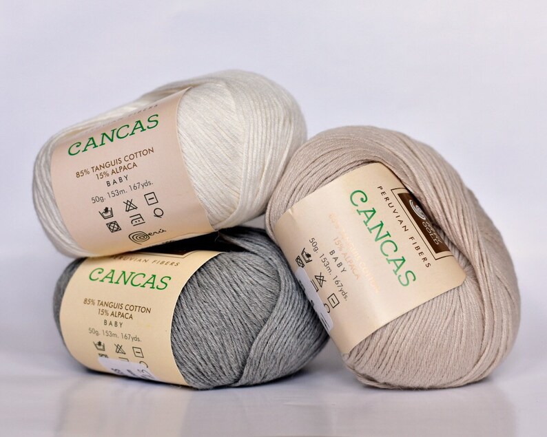 May include: Three skeins of yarn in white, beige, and gray. Each skein has a label that reads "CANCAS" and includes the yarn composition: 85% Tangus Cotton and 15% Alpaca. The label also states "Baby" and the weight, length, and yardage of the yarn.