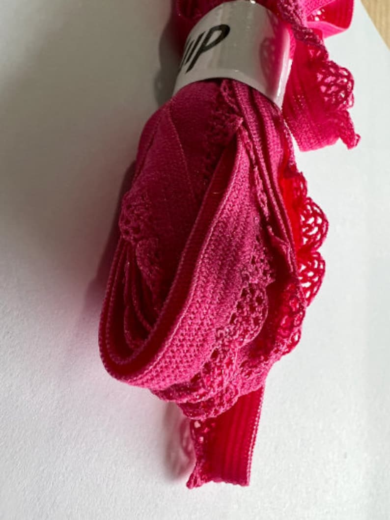 May include: Pink elastic trim with a lace design. The trim is rolled up and secured with a white label that says "Flip".