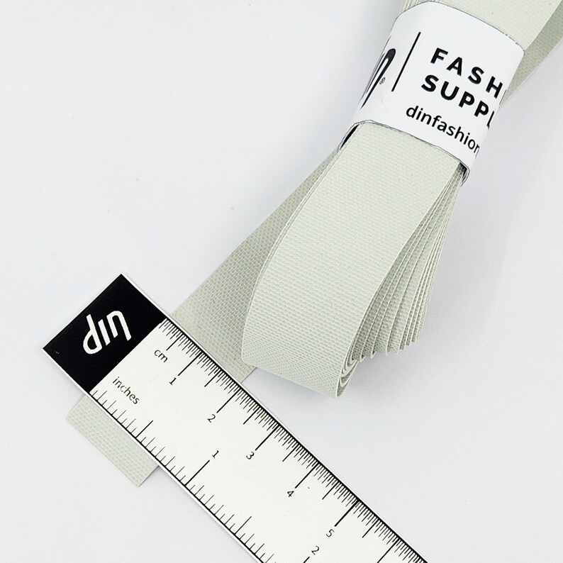 May include: A roll of light grey elastic ribbon measuring 12.7 cm long. The ribbon is folded and has a white label with the text "FASH SUPP dinfashion".