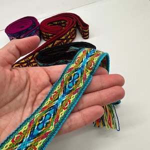 May include: A close-up of a hand holding a bright blue, green, yellow, and red woven ribbon with a geometric pattern. The ribbon has a fringe on one end.