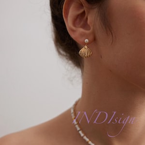 May include: Gold seashell earrings with a pearl accent. The earrings are worn by a person with a white beaded necklace.