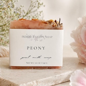 Puede incluir: Una pastilla rectangular de jabón de leche de cabra con peonía y una etiqueta blanca. La etiqueta dice "Indigo Canyon Soap" y "Peony". El jabón está cubierto con plantas secas. El peso neto es de 141 g. Una flor de peonía rosa y ramitas de flores blancas están en el fondo.