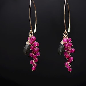 Peut inclure: Une paire de boucles d'oreilles pendantes avec de longs crochets dorés incurvés. Chaque boucle présente une grappe de petites perles facettées rose fuchsia vif et une pierre unique plus grande en forme de goutte gris foncé sur fil doré.