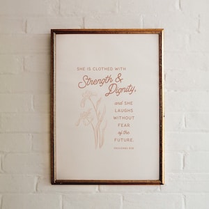 May include: A framed print with a gold-coloured frame. The print displays the quote "She is clothed with Strength & Dignity, and she laughs without fear of the future. Proverbs 31:25" in a warm tone, accompanied by a floral illustration.