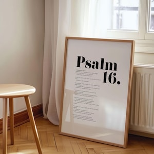 May include: A framed print displaying the text "Psalm 16" in bold black lettering. The print contains the full text of Psalm 16 in a smaller font. The frame is light brown. A small wooden stool is in the foreground.